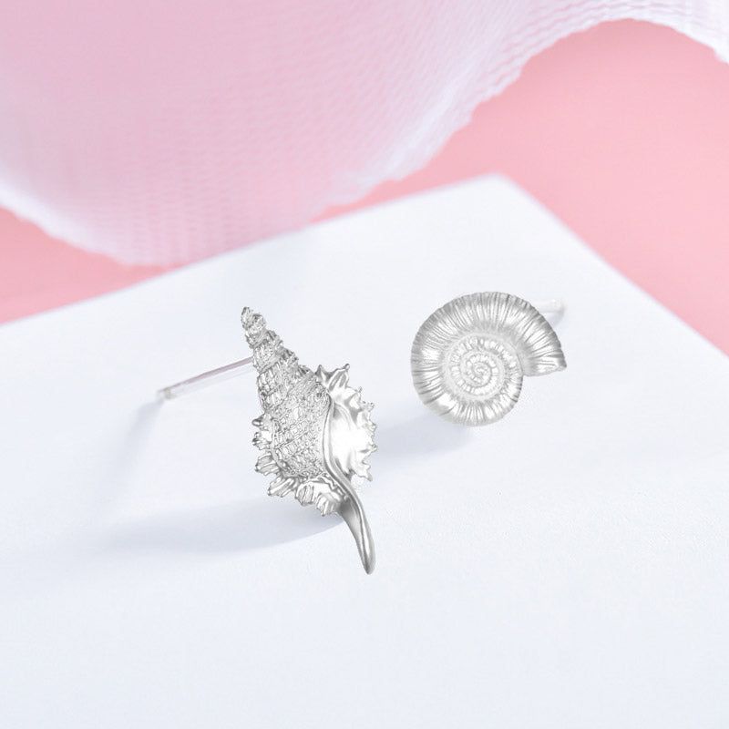 A set of five pairs of 925 sterling silver asymmetrical sea shell and conch stud earrings displayed on a pink and white background.