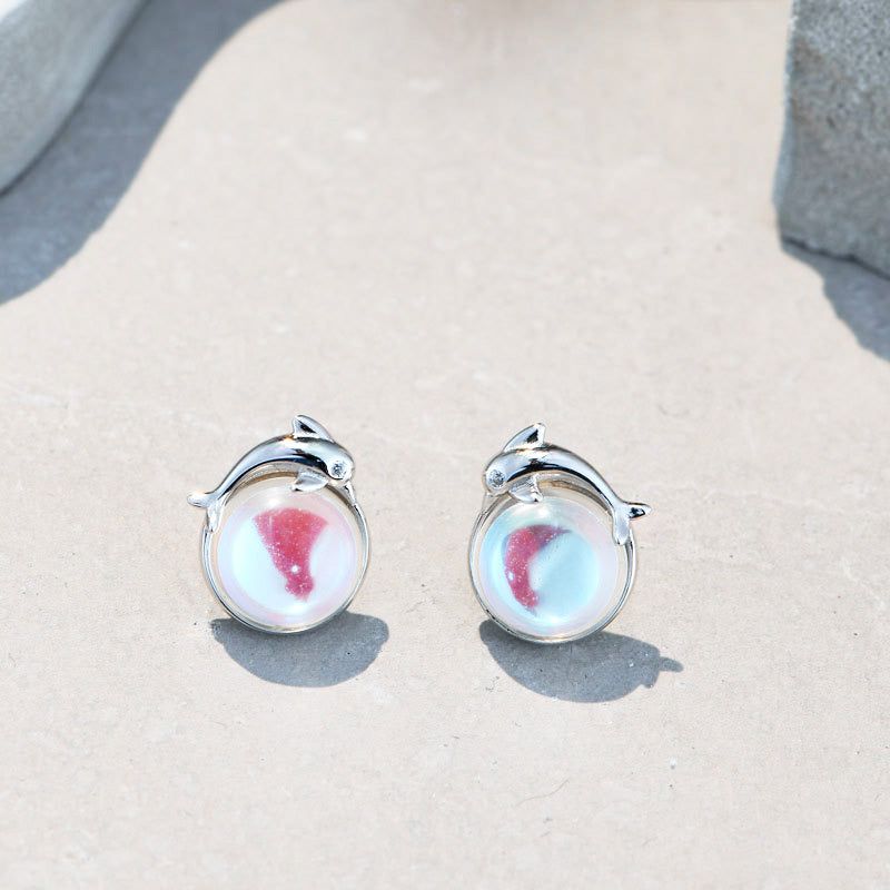 Un primo piano degli orecchini a bottone in argento sterling 925 con pietra di luna a forma di delfino, che mette in mostra il dettagliato montaggio in argento a forma di delfino e la pietra di luna iridescente.