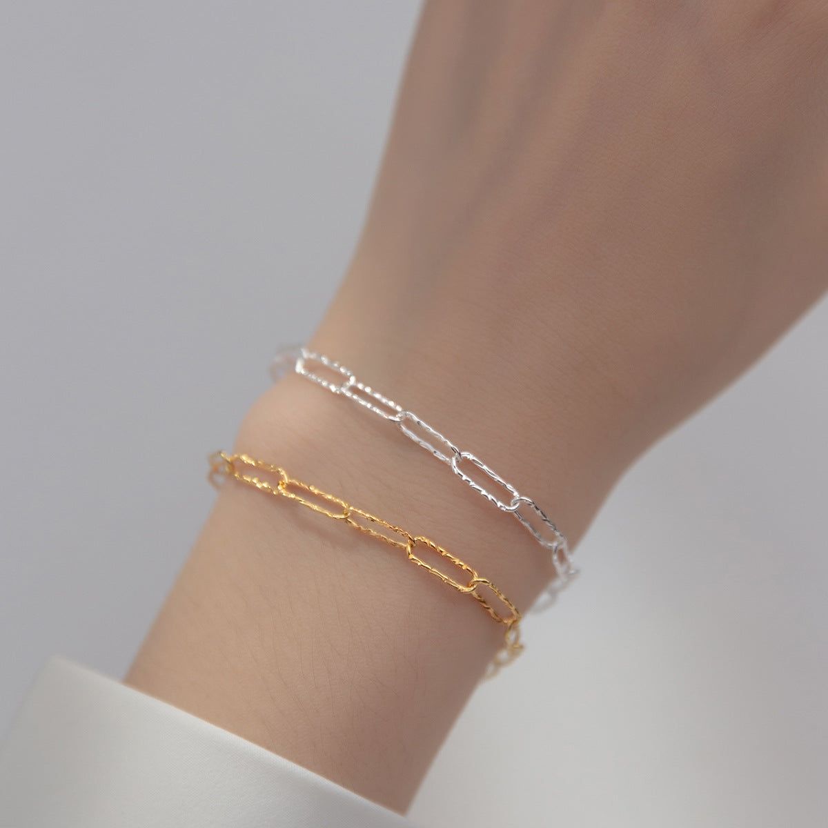 Um conjunto de pulseiras de corrente de clipe de papel de prata esterlina 925 em acabamentos prateados e dourados, elegantemente usadas em um pulso para mostrar o design cintilante.