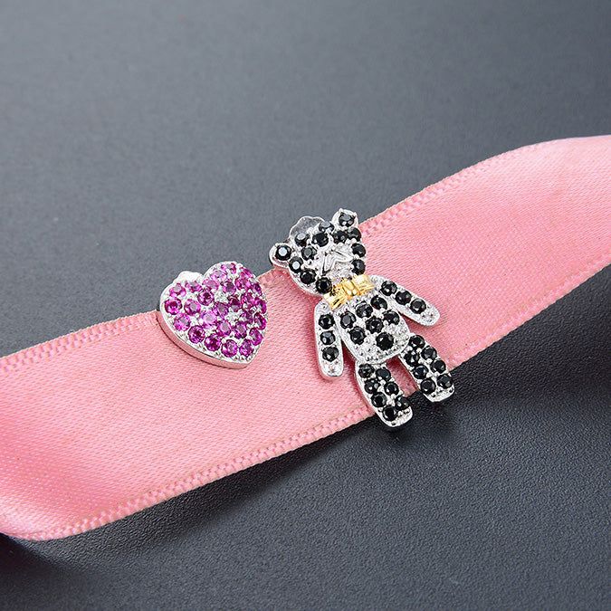 Un affascinante set di orecchini a perno in argento sterling 925 con un cuore di strass rosa e un design di orsetto di strass neri, esposti su un nastro rosa.