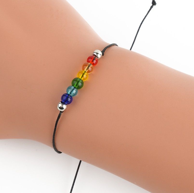 A close-up of the handmade rainbow LGBT rope bracelet being worn, highlighting its colorful acrylic beads and adjustable black cord.