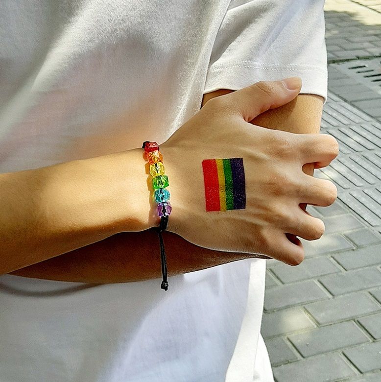 Il polso di un uomo che indossa il Braccialetto Arcobaleno per Uomini, con perline acriliche vivaci, insieme a un tatuaggio temporaneo arcobaleno abbinato, simbolo del orgoglio LGBTQ+.
