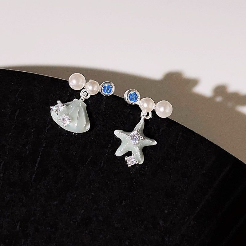 星の砂や貝殻のデザインに真珠と青いジルコンのアクセントが施された、海をテーマにした銀メッキのスタッドピアス5ペアのコレクションが白い表面に展示されています。