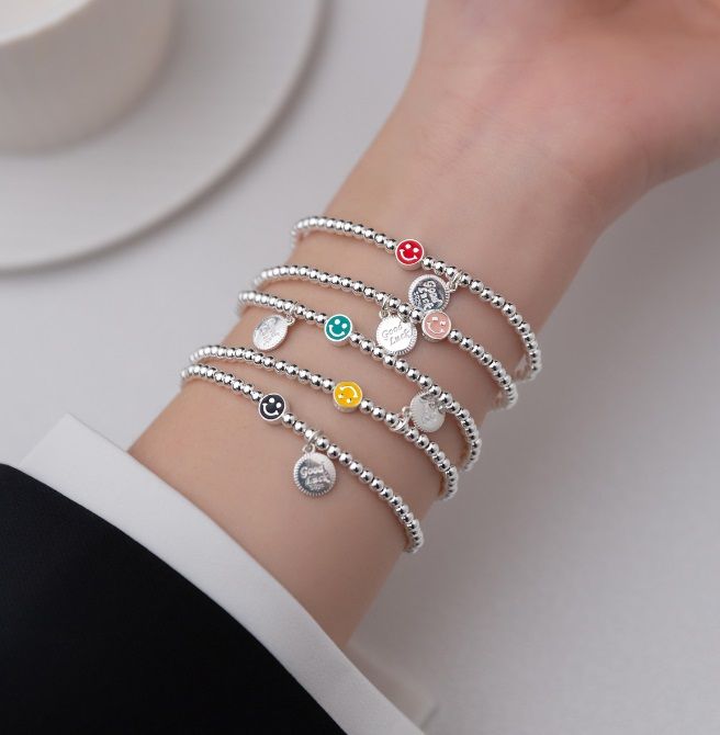 Una collezione di braccialetti elastici in argento sterling 925 con faccine sorridenti e ciondoli smaltati colorati, esposti su uno sfondo neutro. Modello JRSH-011 di JR Fashion Accessories.