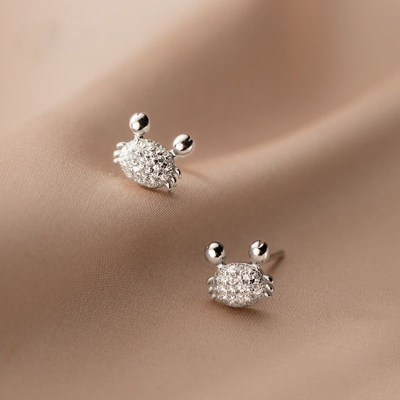 Un paio di orecchini a bottone a forma di granchio in argento sterling 925, dettagliati in modo intricato con scintillanti pietre di zirconia cubica, esposti su uno sfondo neutro.