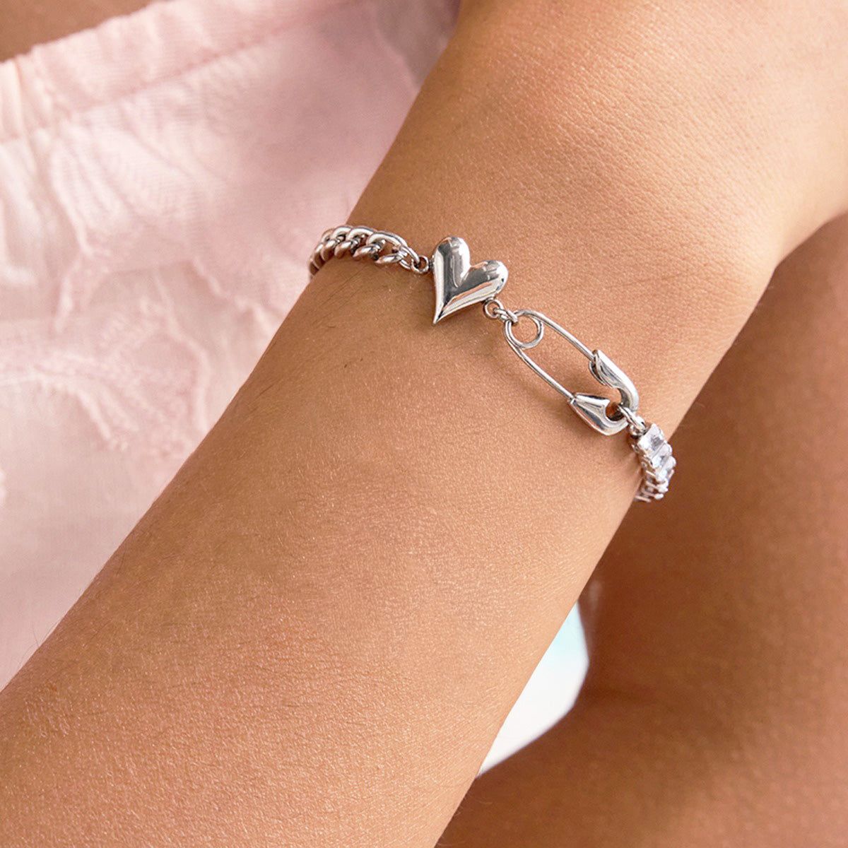 Ett tennisarmband i rostfritt stål med hjärtcharm och säkerhetsnålsdesign, prytt med kubisk zirkonia, elegant bärs på en handled. Del av ett 5-delars set från JR Fashion Accessories.
