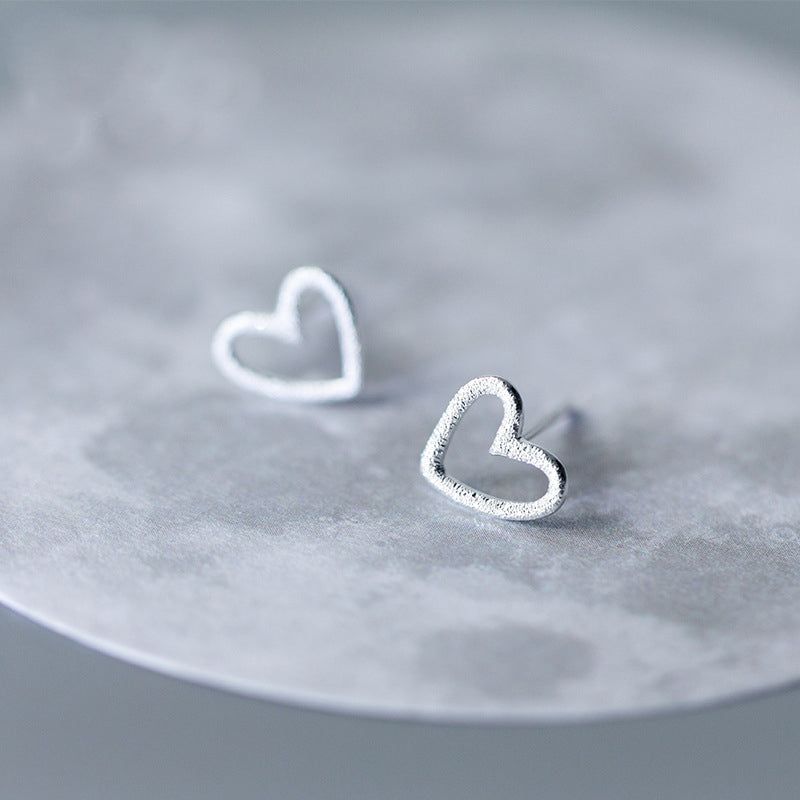 Un paio di orecchini a perno a forma di cuore vuoto in argento sterling 925 con una finitura testurizzata, esposti su uno sfondo bianco pulito.
