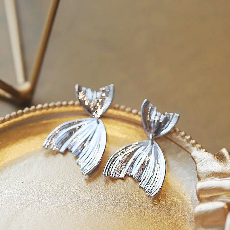 Ein Paar hängende Ohrringe in Form eines Meerjungfrauen-Schwanzes aus 925er Sterlingsilber, elegant auf einem goldenen Teller präsentiert, um ihr filigranes Design zu zeigen.