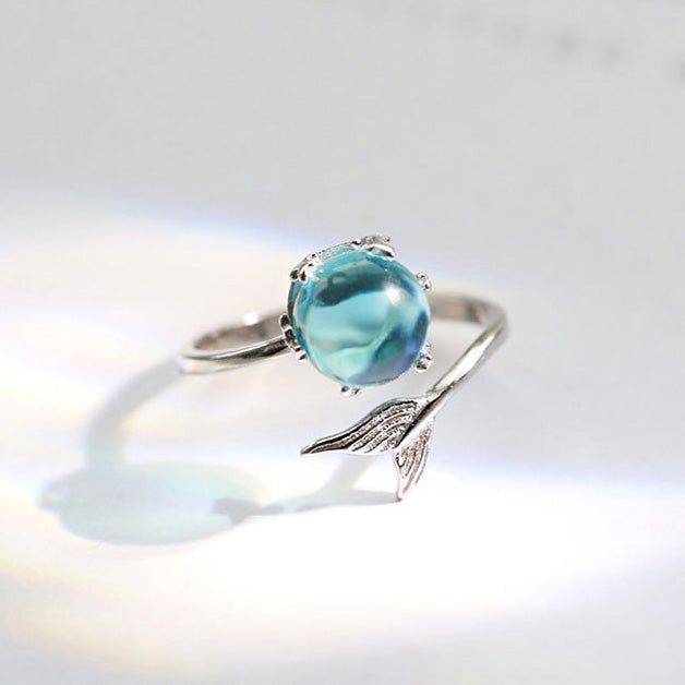Eine Nahaufnahme eines 925er Sterlingsilberrings aus dem Meerjungfrauen-Schwanz-Motiv-Set, der ein filigranes Flügel-Design und einen funkelnden hellblauen Edelstein zeigt.