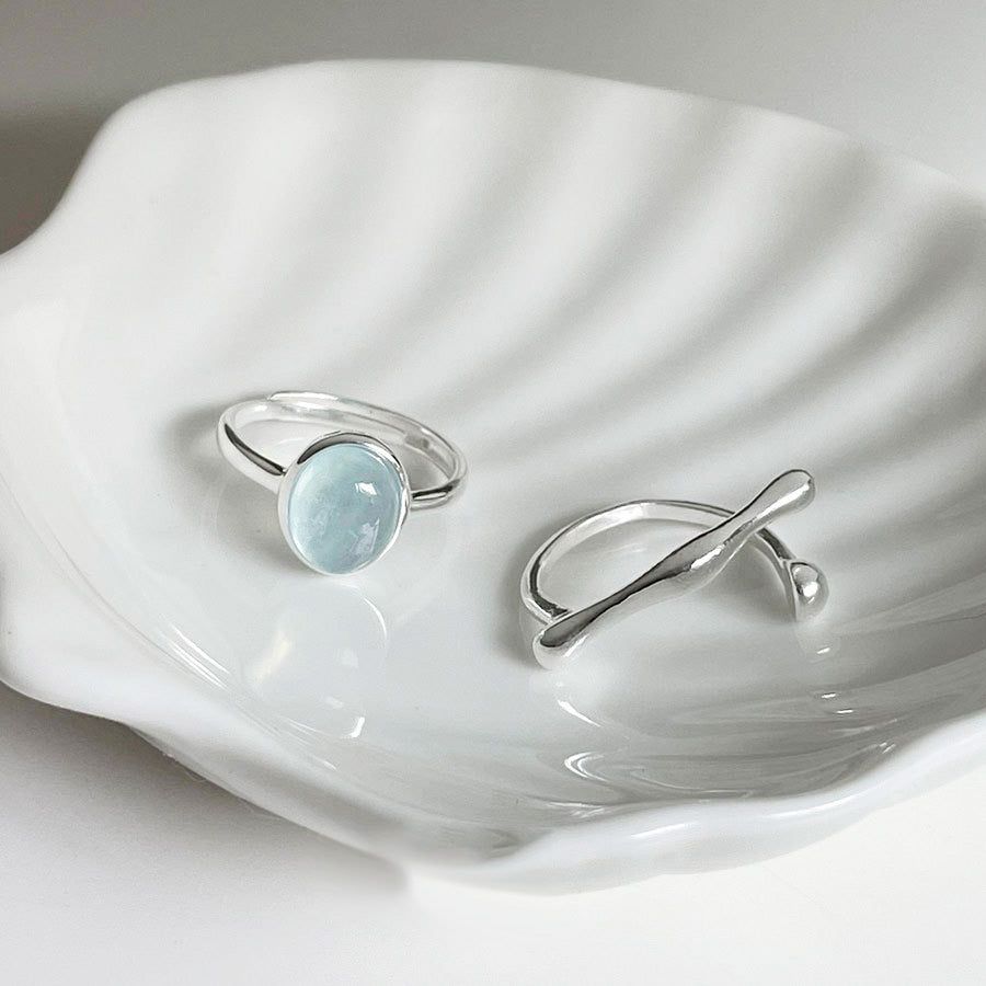 Un conjunto de anillos ajustables de plata esterlina 925, uno con una piedra azul claro y otro con una banda retorcida, exhibidos en un plato decorativo.