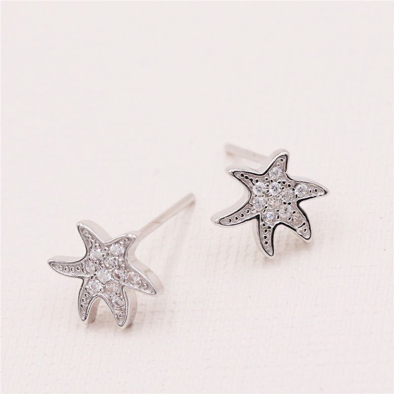 Un paio di orecchini a forma di stella marina in argento sterling 925, dettagliati in modo intricato con piccole gemme trasparenti, presentati su uno sfondo bianco pulito.