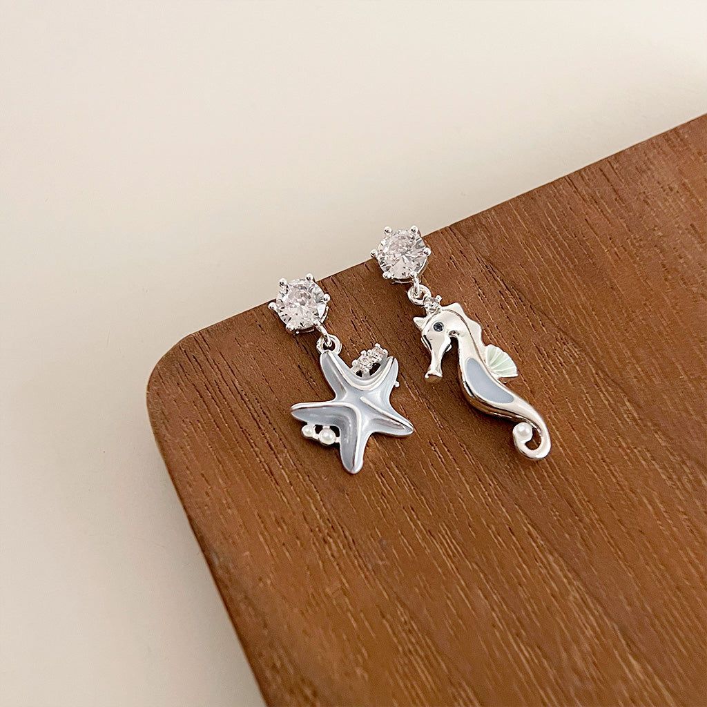 Un paio di orecchini a goccia asimmetrici a tema oceanico con una stella marina in argento e un cavalluccio marino, entrambi accentuati con pietre trasparenti, su uno sfondo bianco pulito.