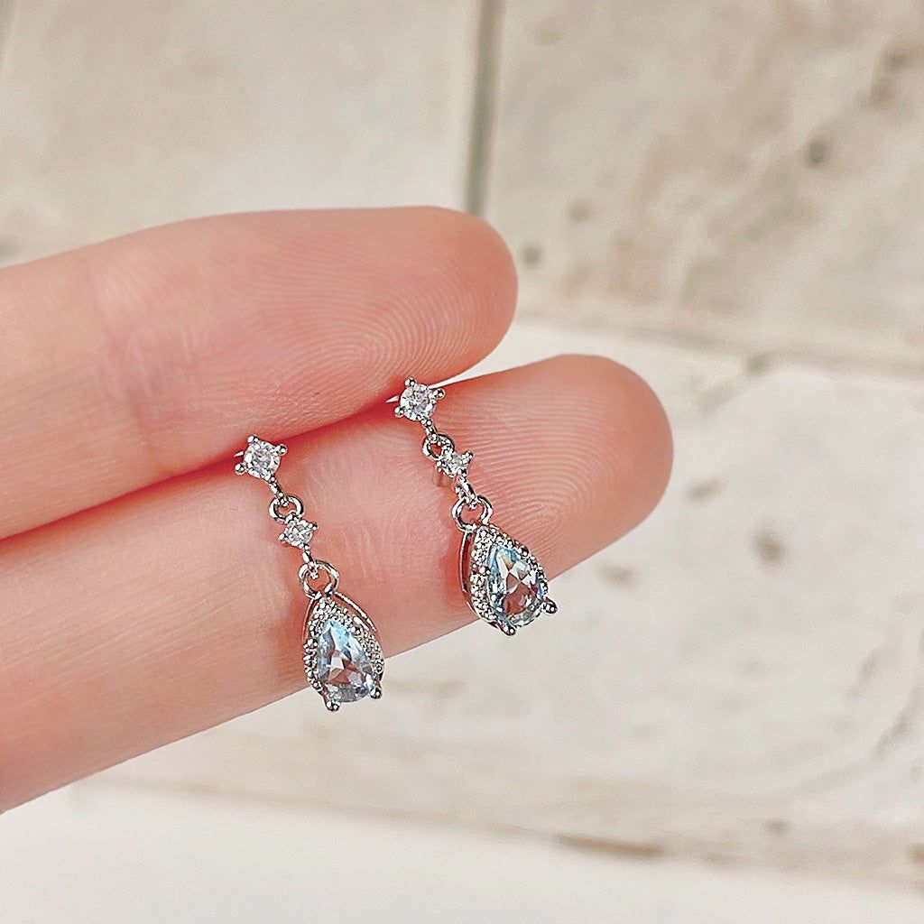 Un primer plano de los pendientes de gota de agua azul de zirconia chapados en plata, mostrando el intrincado diseño con una gran piedra en forma de lágrima rodeada de piedras claras más pequeñas.