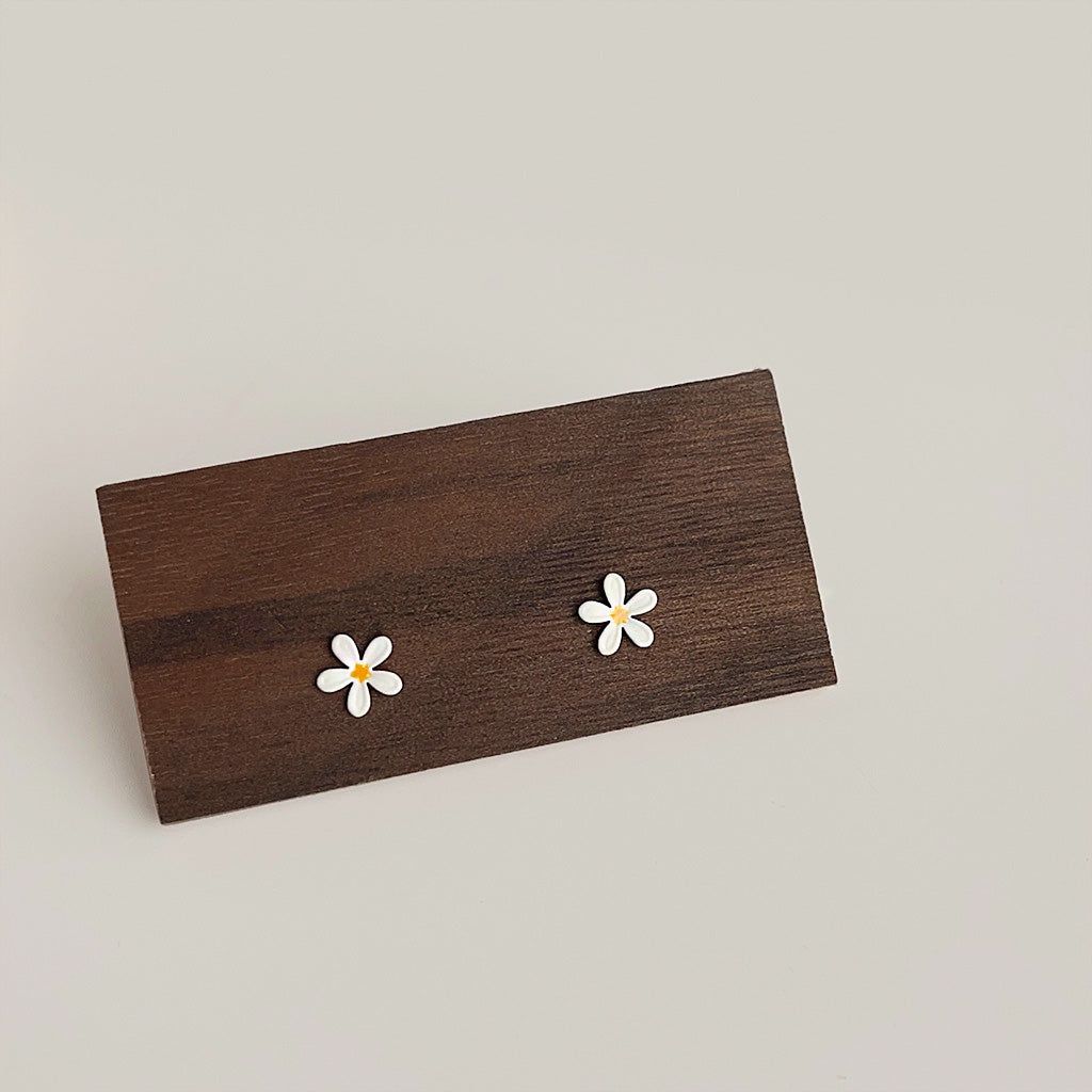 Un par de pendientes de botón de mini flor de margarita de esmalte, adorables y al por mayor, exhibidos sobre un bloque de madera rústica, destacando sus pétalos blancos y centros amarillos.