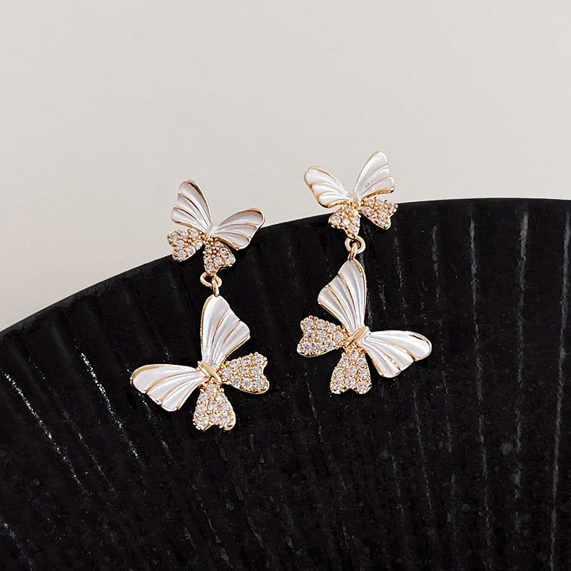 A pair of 14k gold-plated statement drop earrings featuring a double white enamel butterfly design with gold trim and sparkling zircon accents.