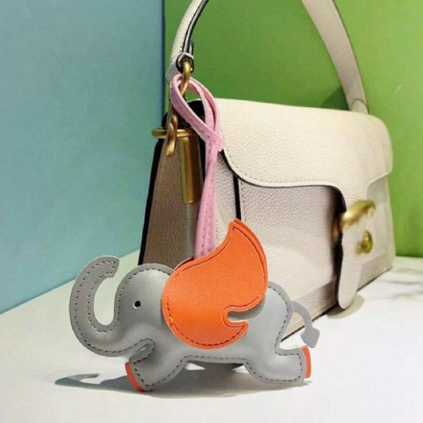 Un lindo charm de bolso en cuero PU de elefante blanco con acentos coloridos, adjunto a la correa de un elegante bolso blanco.
