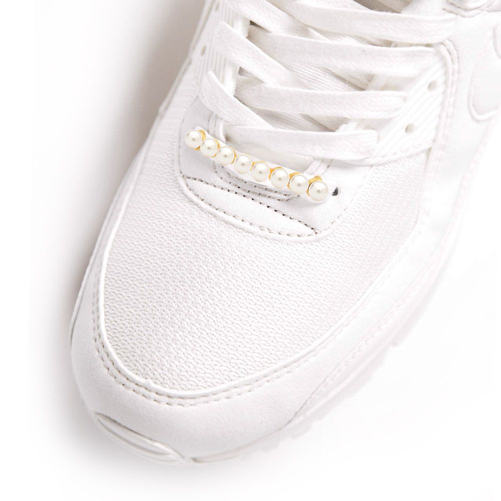 Primer plano de un encantador de zapato de perla elegante con chapado en oro y perlas blancas adornando los cordones de una zapatilla blanca.