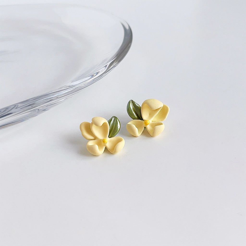 Une paire de boucles d'oreilles en fleur de gardénia 3D en émail jaune avec des détails de feuilles vertes, présentée sur un fond blanc.