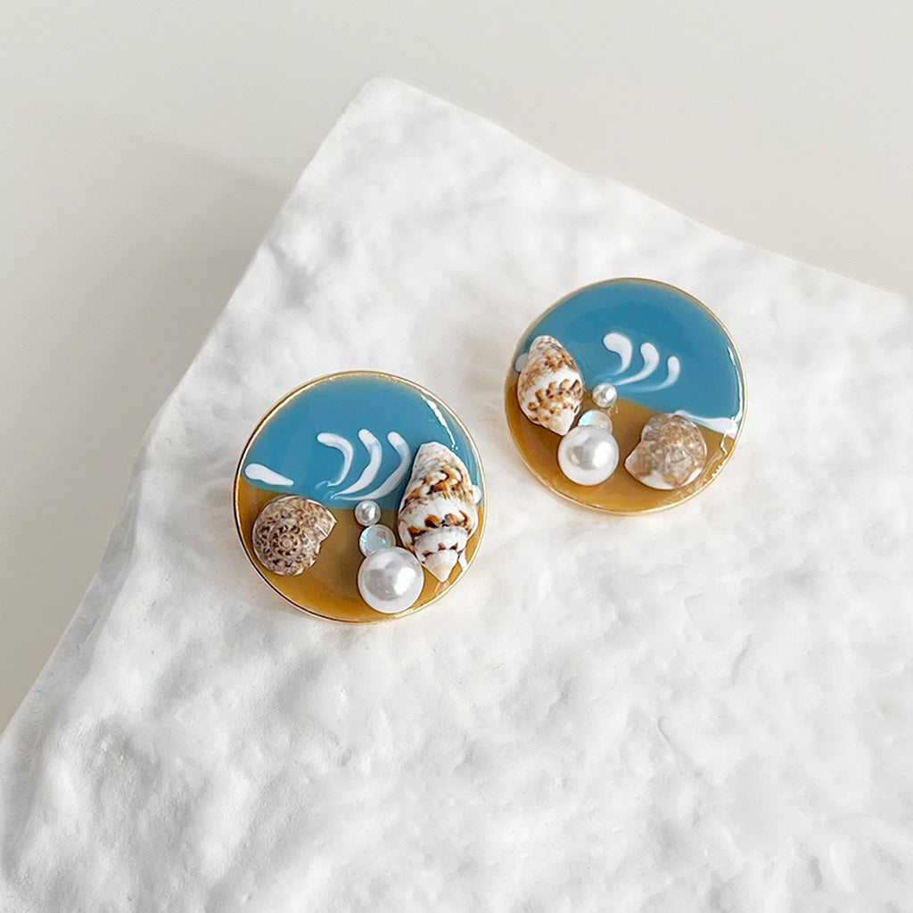 A set of five pairs of summer style beach-themed stud earrings with seashells, pearls, and gold plating, displayed on a sandy background.