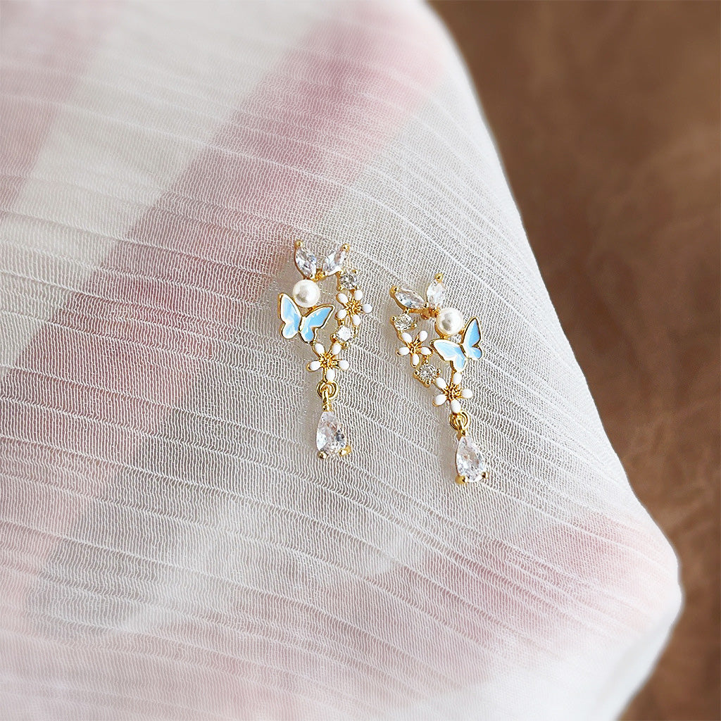 A pair of 14k gold plated earrings featuring a detailed blue enamel butterfly and white flower, accented with a dangling pearl drop.