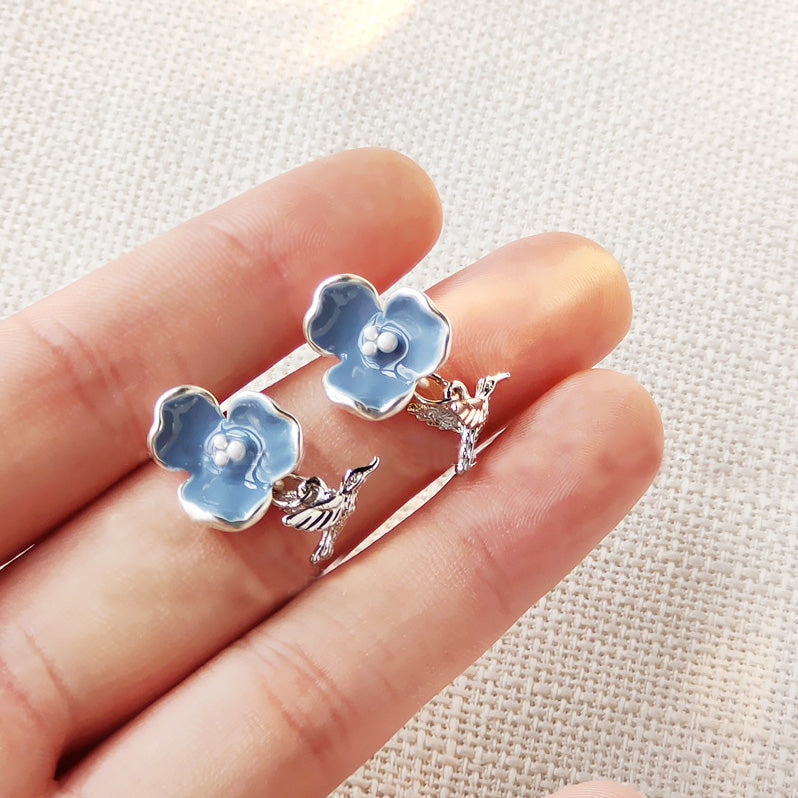 A set of vintage silver-plated stud earrings featuring blue enamel flowers and dangling bird charms, displayed on a neutral background.