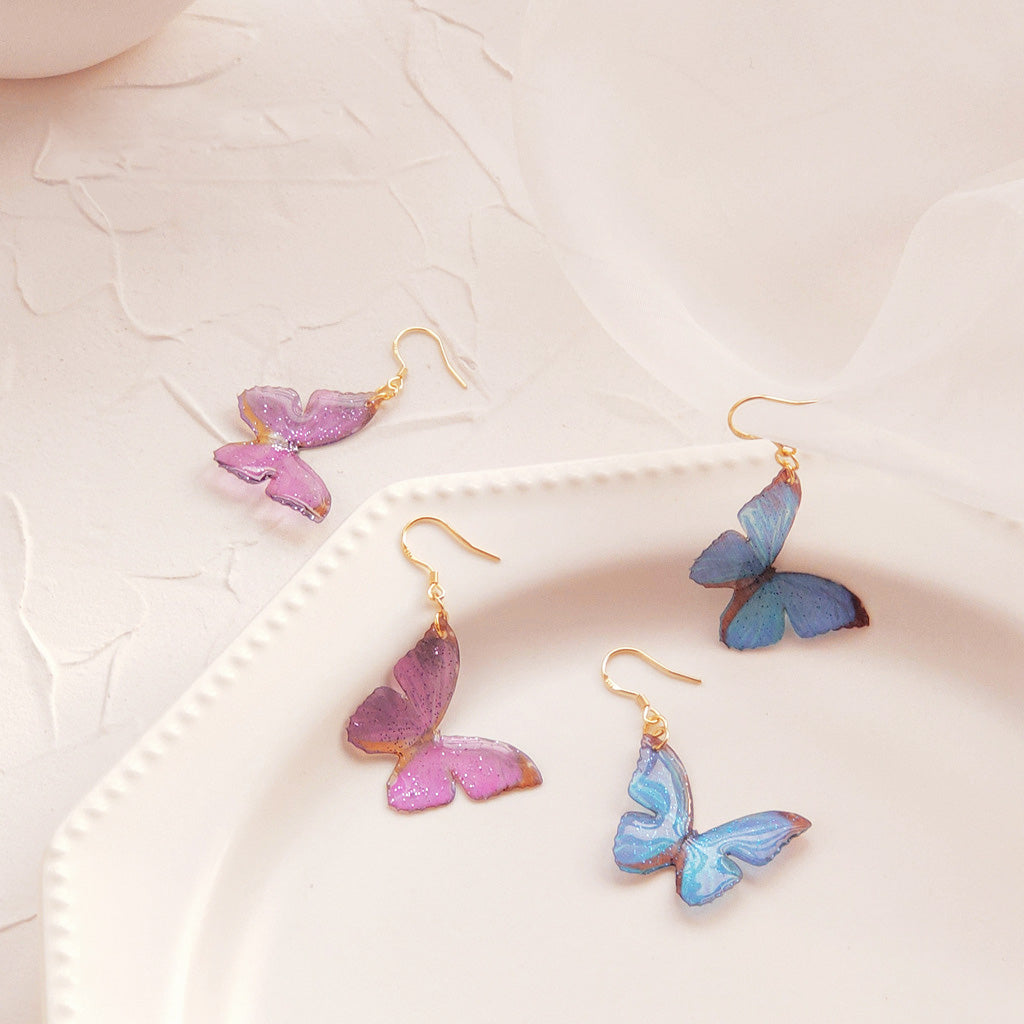 A set of lightweight brass butterfly dangle earrings shown in vibrant purple and blue glittery finishes, with gold-tone hooks.