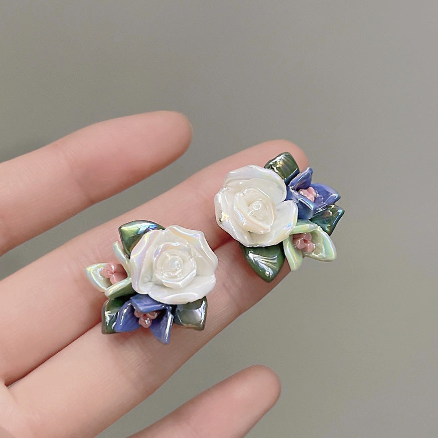 A close-up of a hand holding a pair of delicate 3D white ceramic flower stud earrings, showcasing their intricate floral design.