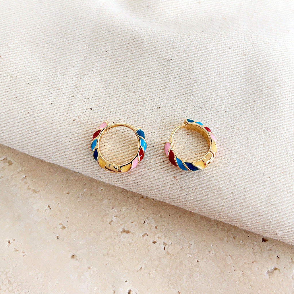 A close-up of a pair of 14k gold-plated huggie hoop earrings with vibrant, multi-colored rainbow enamel stripes.