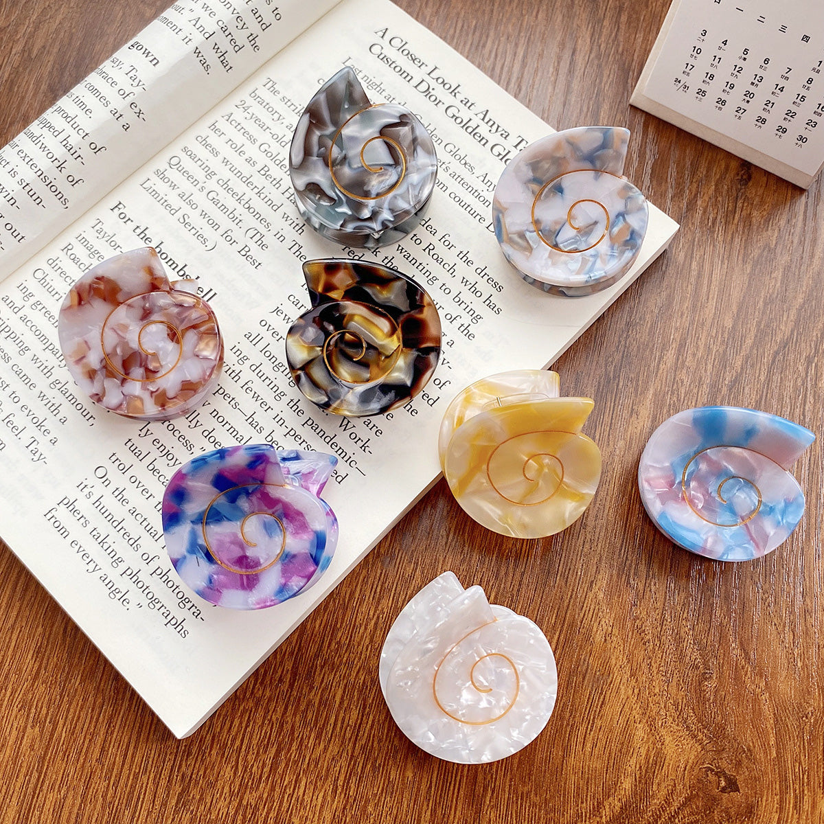 A vibrant collection of small conch shell-shaped acetate hair claw clips in various marbled colors, displayed on a wooden surface.