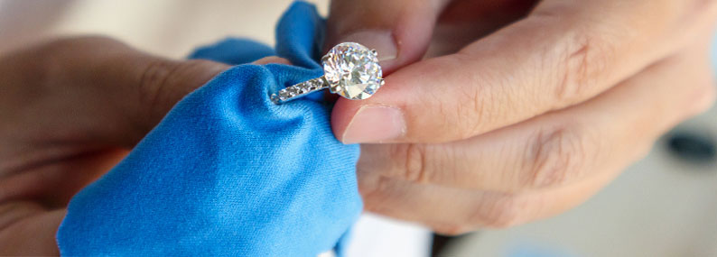 A close-up of a large diamond ring being carefully cleaned with a soft blue cloth, demonstrating proper jewelry care.