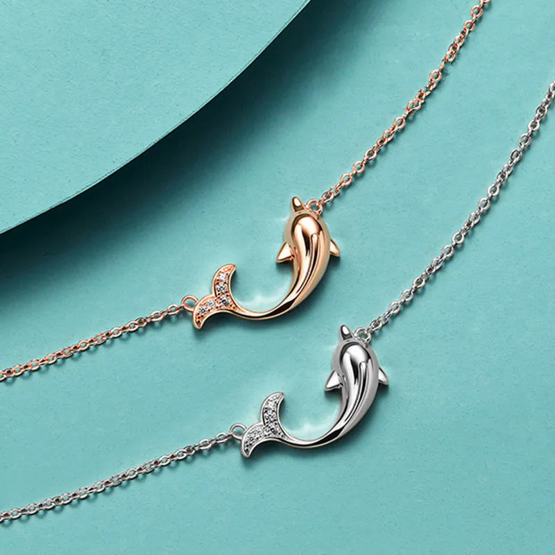 A pair of 925 sterling silver dolphin pendant necklaces, one in a polished silver finish and the other in a warm rose gold finish, both featuring sparkling stone-studded tails.