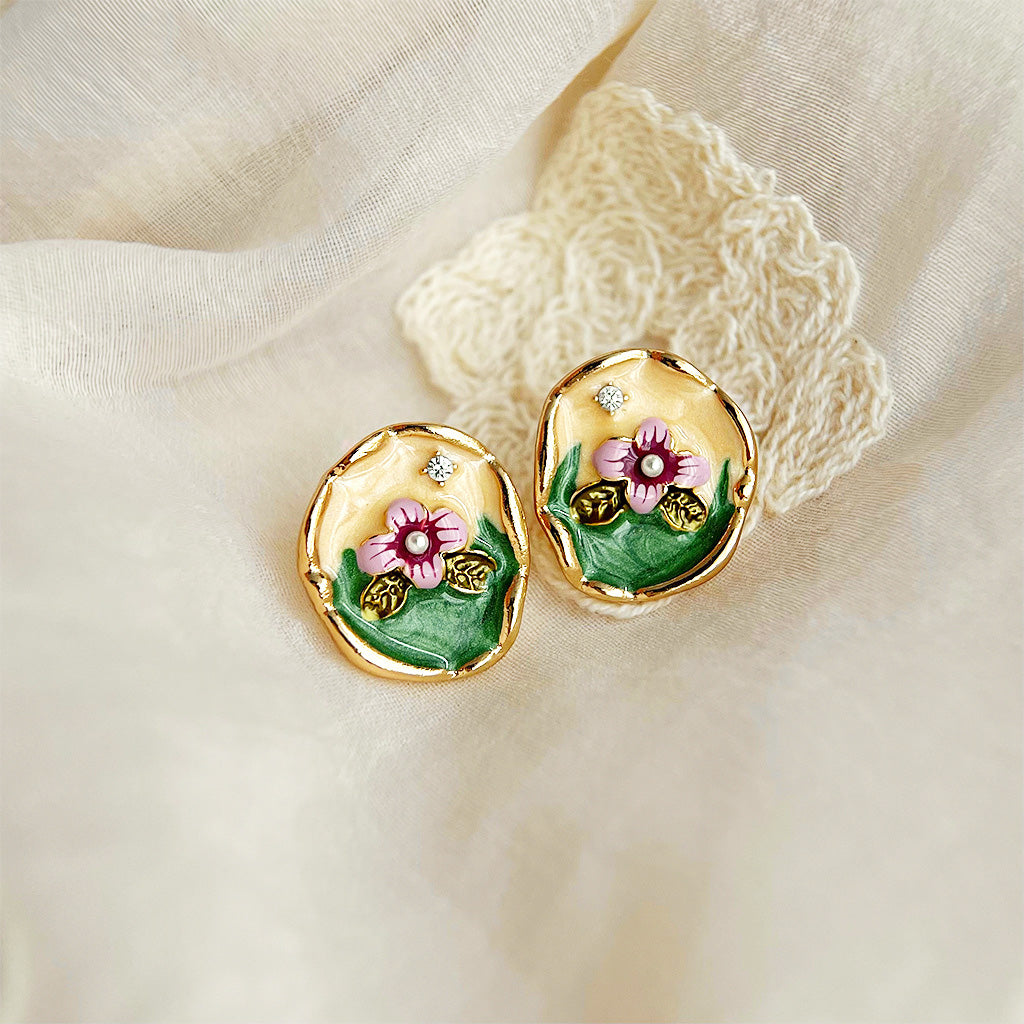 A pair of 14k gold-plated floral stud earrings featuring delicate pink enamel flowers, green leaves, and accented with shimmering rhinestones and a central pearl.