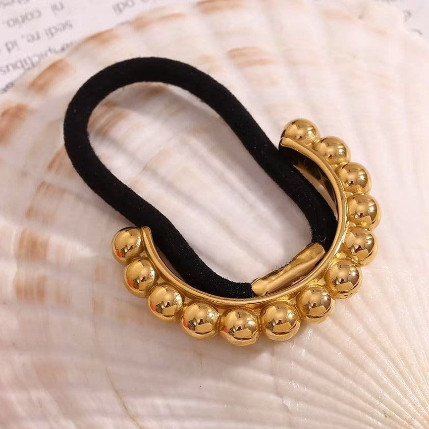 A stylish 18K gold plated stainless steel hair cuff with a black elastic tie, featuring a decorative gold bead detail, displayed on a seashell.