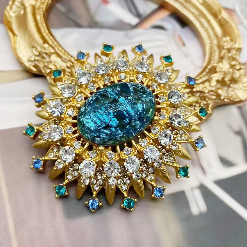 A luxurious gold-plated vintage sun brooch pin with a large central blue gemstone surrounded by sparkling clear and blue crystals.