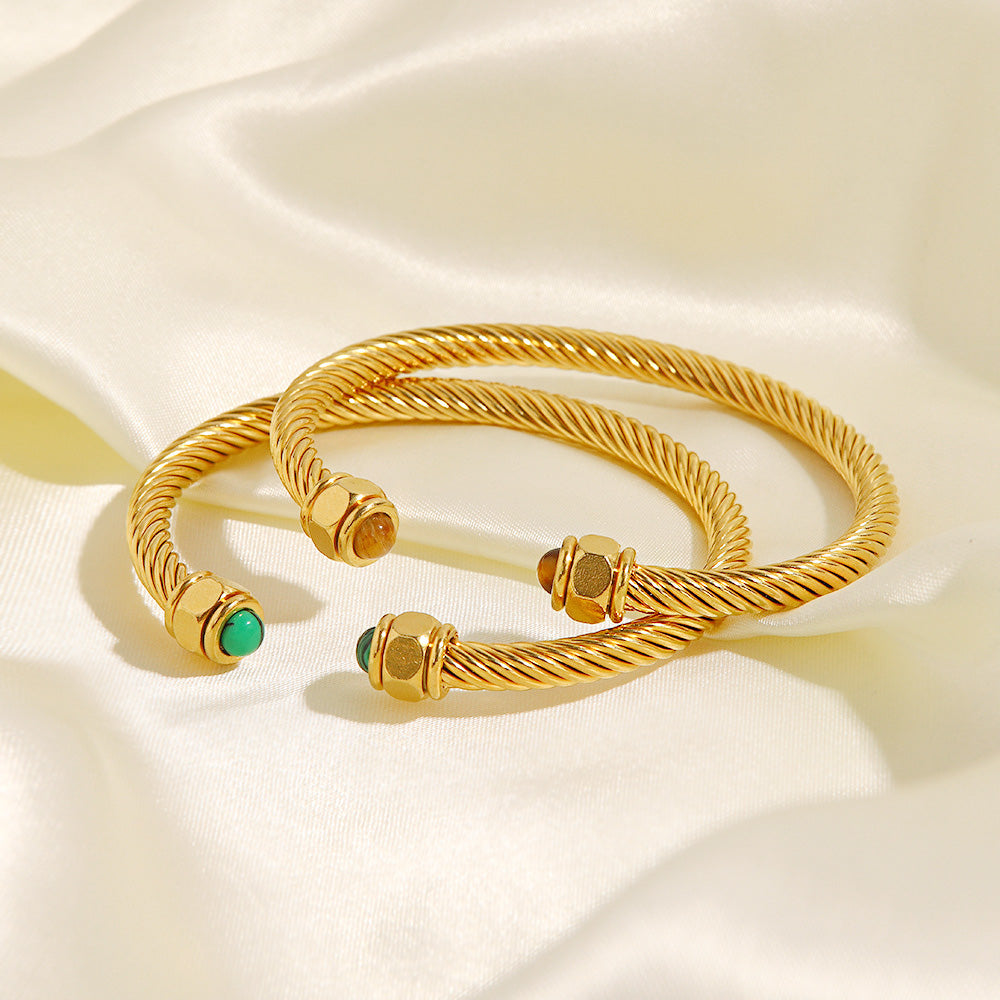 A set of two stainless steel open cuff bangles with a twisted design, one with green and one with brown natural stone accents, displayed on a neutral background.