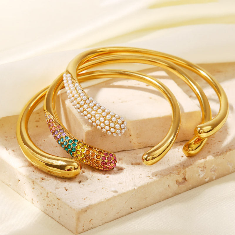 A set of five stainless steel gold-tone open cuff bangles, featuring designs with colorful cubic zirconia and elegant pearls.