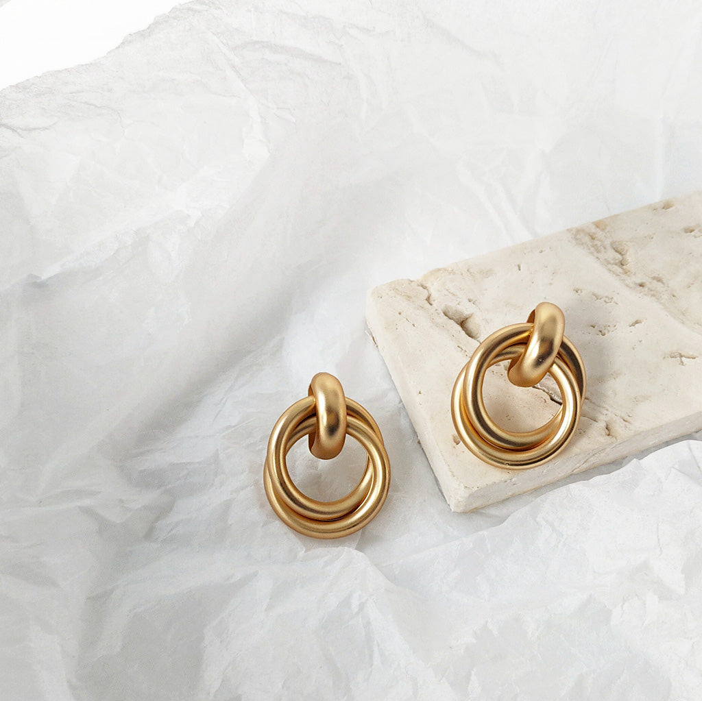A pair of matte gold-plated geometric knot cord stud earrings with a distinctive double-ring design, on a clean white background.