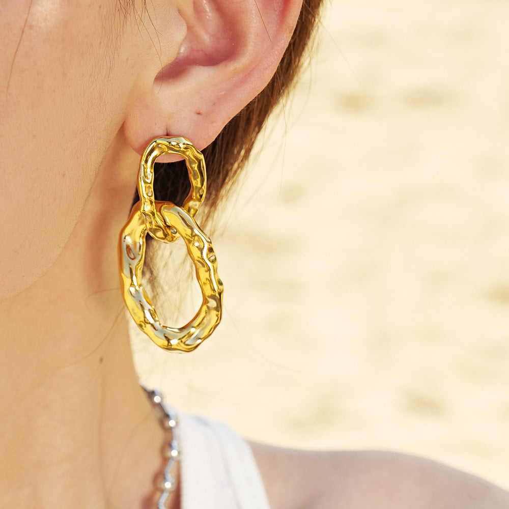 Close-up of a chunky gold elliptical interlock charm drop earring made from stainless steel, showcasing its textured finish.