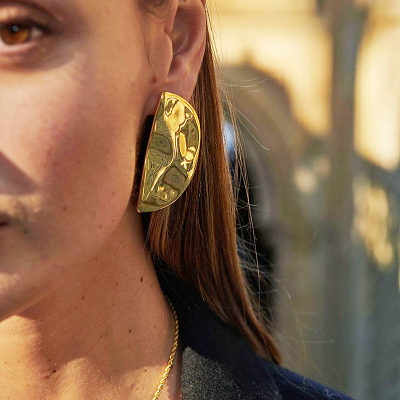 A close-up view of a woman wearing a gold stainless steel half-circle stud earring with a shiny hammered finish, showcasing its texture and design.