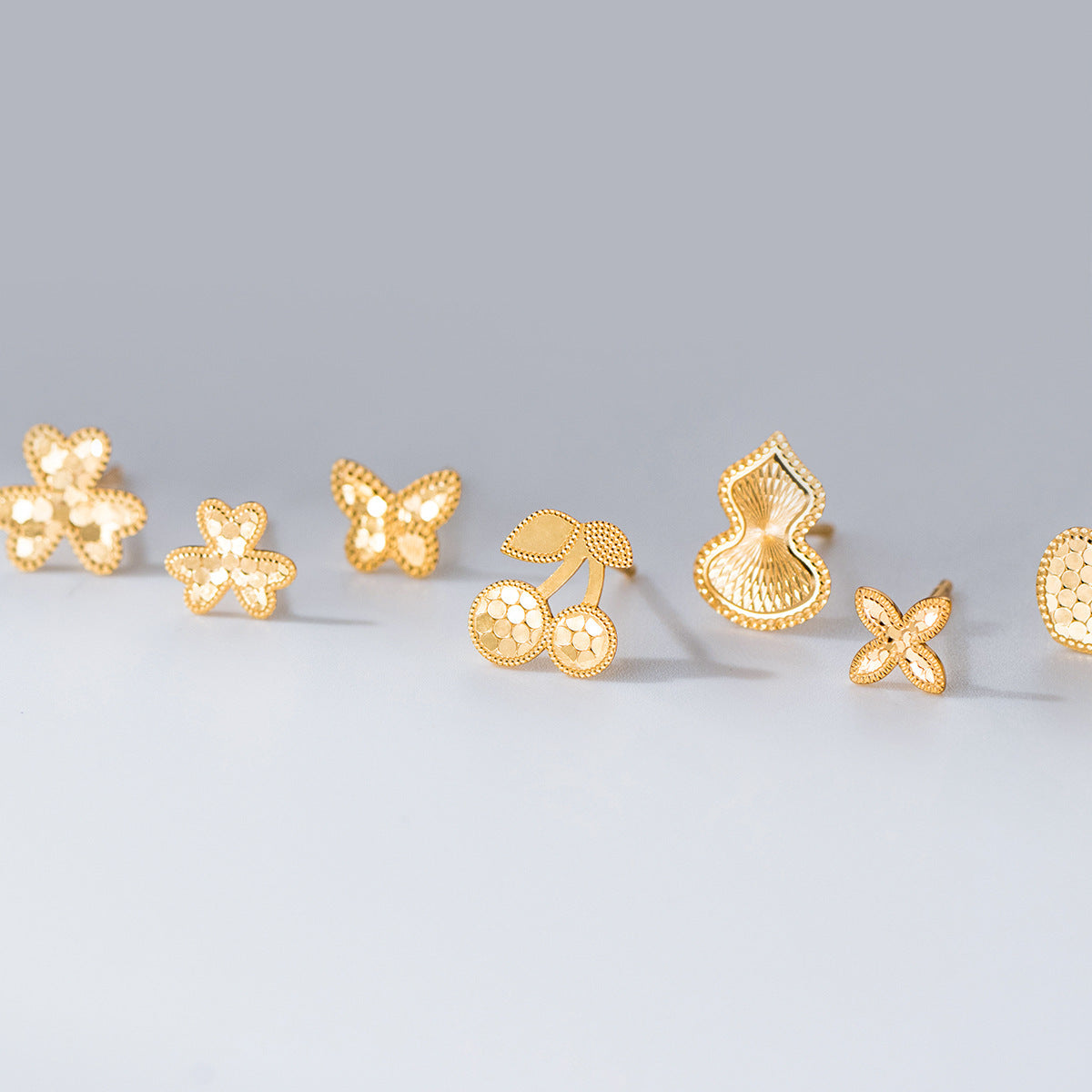 A collection of five pairs of wholesale 925 sterling silver laser-cut stud earrings in gold, featuring flower, butterfly, heart, cherry, and gourd designs, arranged on a white background.