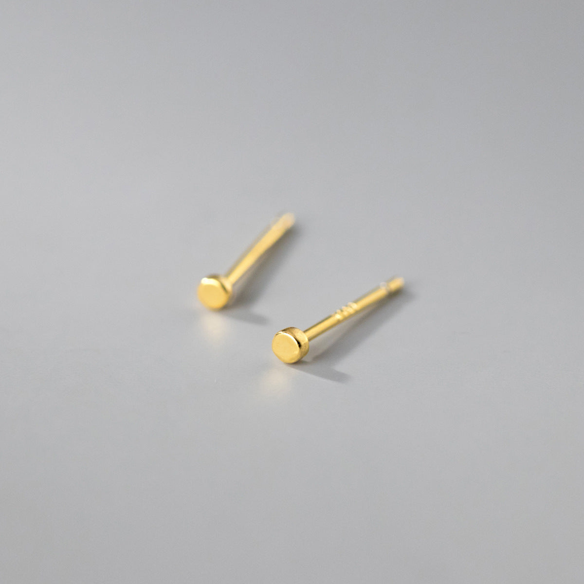 A pair of simple, tiny circle stud earrings in a polished gold finish, made from 925 sterling silver, displayed on a white background.