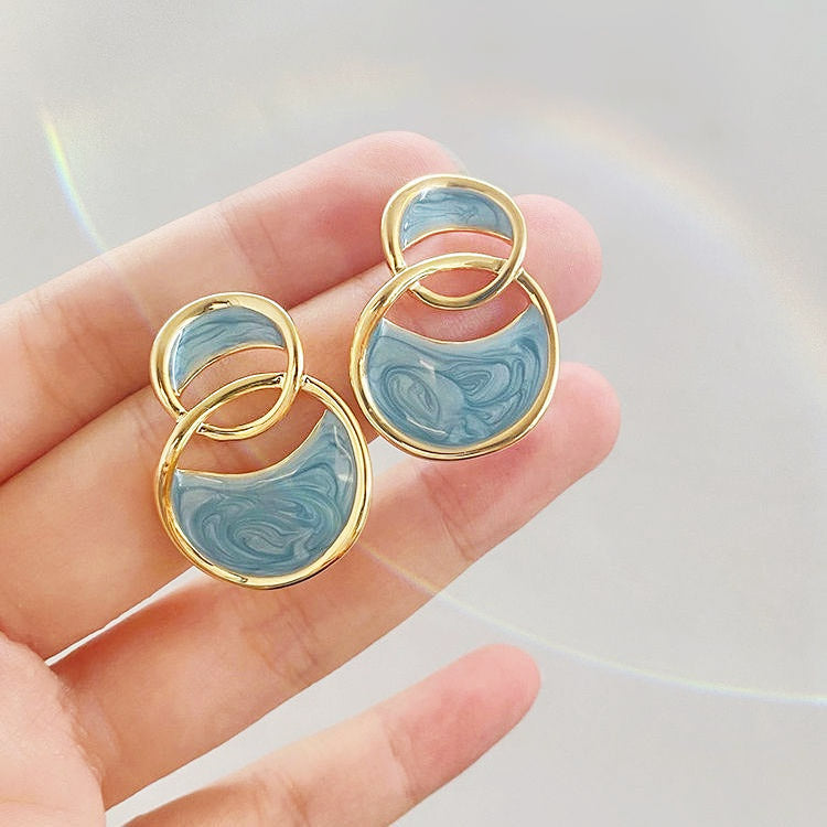 A pair of hollow-out 14k gold plated interlocking circle stud earrings with a vibrant blue and white marbled enamel inlay, shown on a white background.