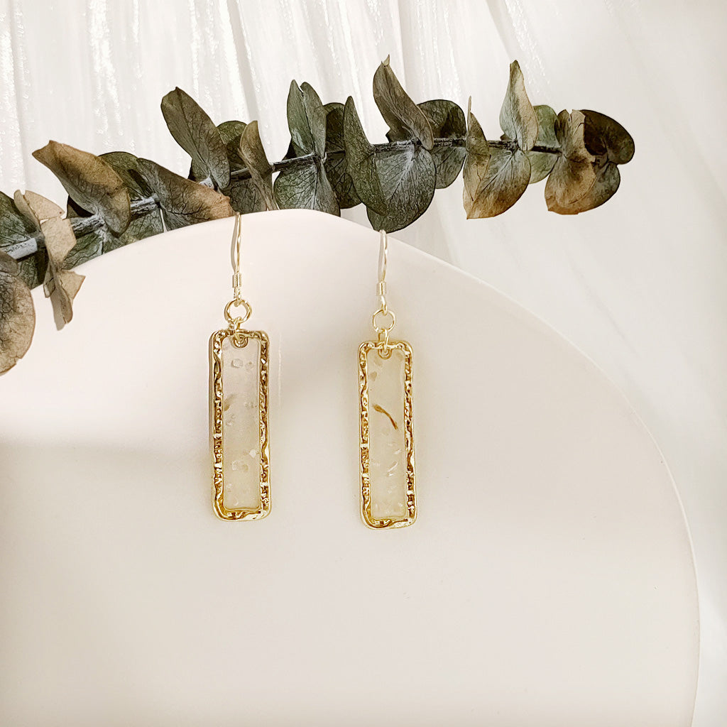 A pair of geometric rectangular gold frame earrings with transparent resin and shell inclusions, styled with eucalyptus leaves on a white surface.