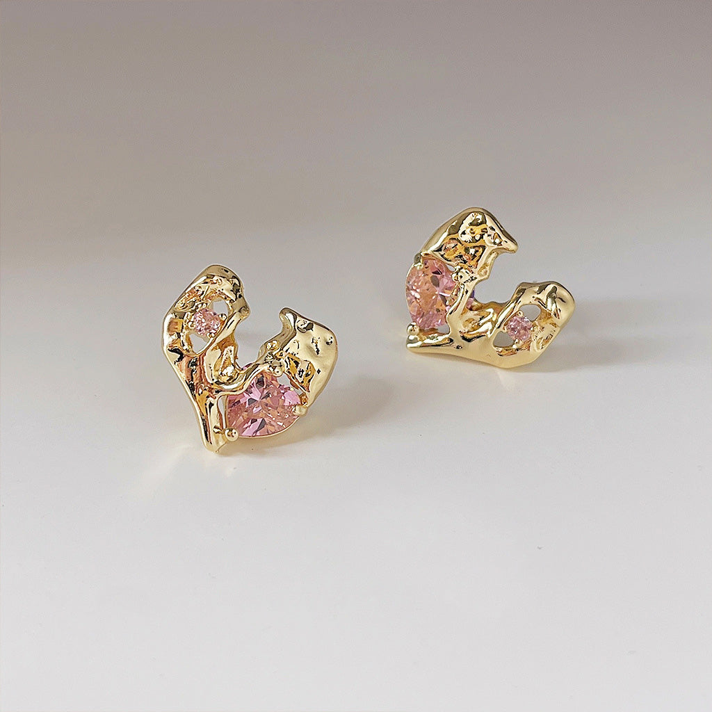 A close-up shot of a pair of gold-plated irregular heart stud earrings, each featuring a vibrant pink crystal gemstone.