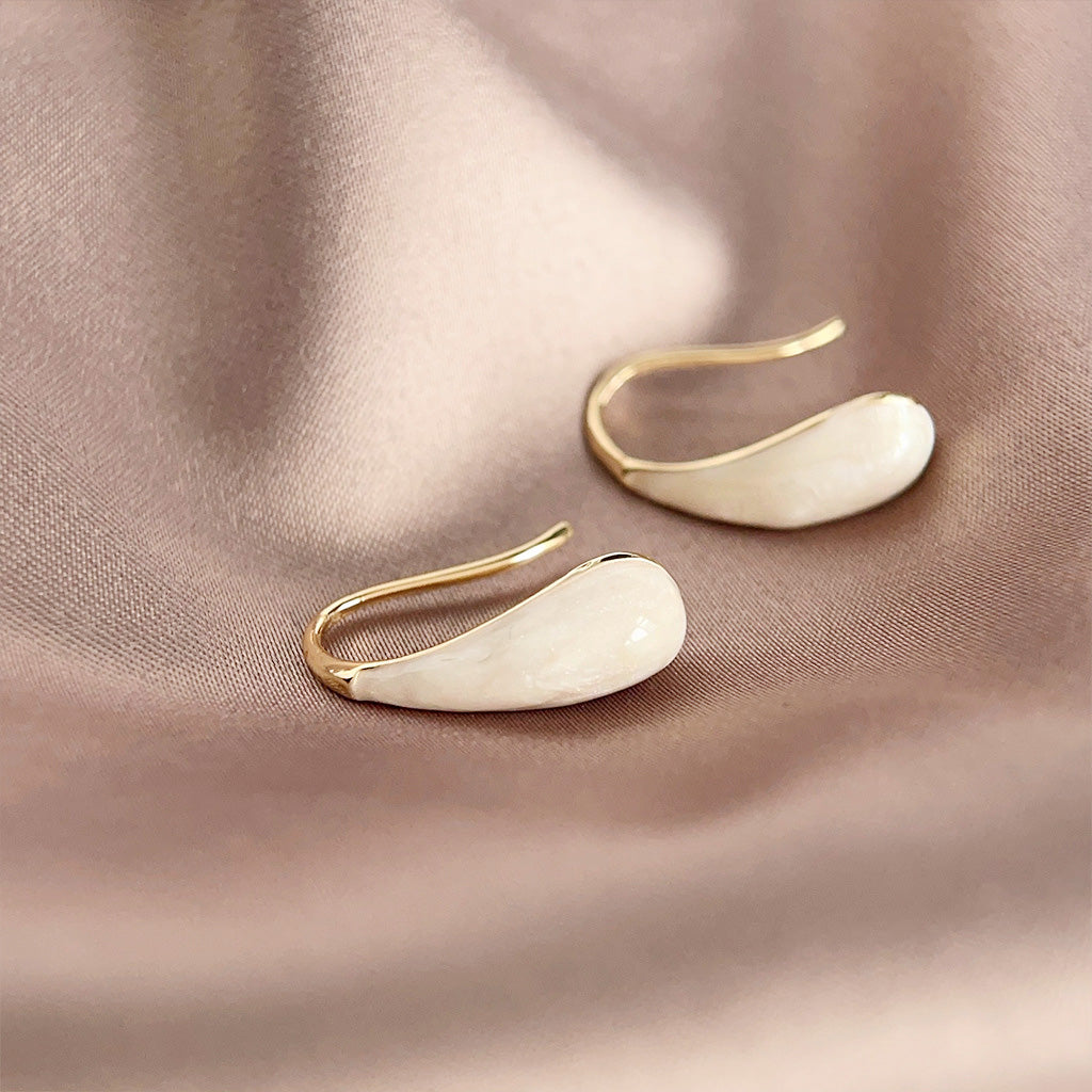 A pair of wholesale gold-plated French hook earrings with white cream enamel waterdrop pendants, displayed on a textured beige surface.