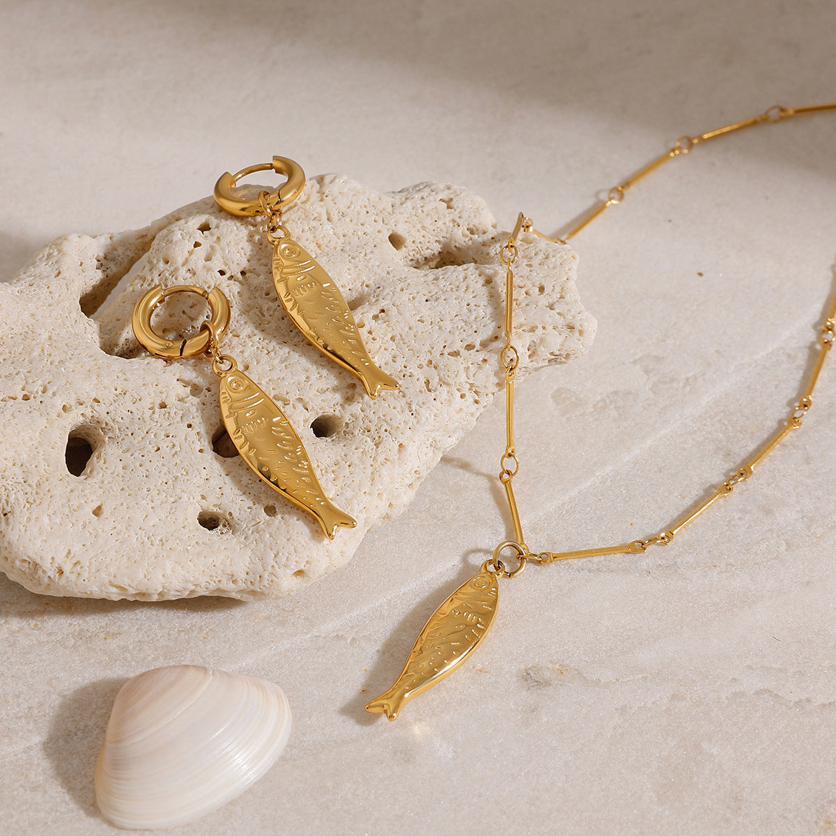 A stunning gold fish-shaped jewelry set, including a necklace and earrings, elegantly displayed on a beige stone with a seashell, perfect for beach-themed styles.
