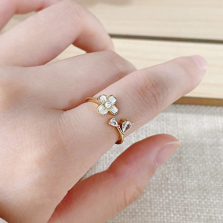 A detailed close-up of a 14k gold-plated brass ring from a set of five, featuring an intricate white enamel flower and butterfly design with zircon accents.