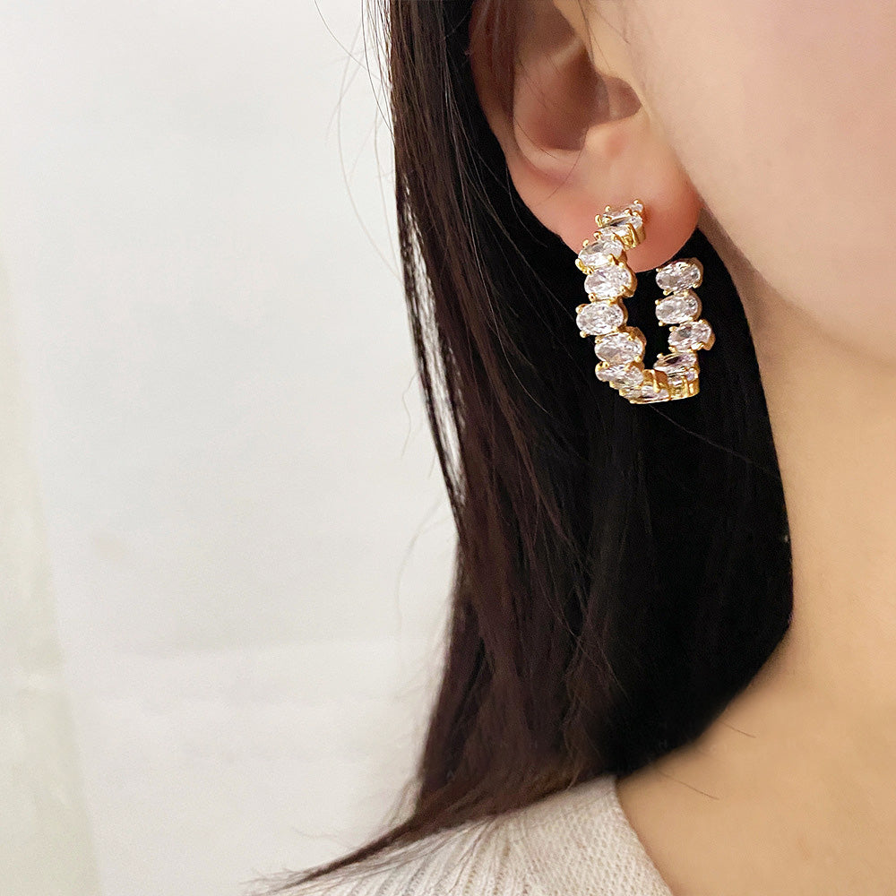 A close-up of a gold-plated C-hoop earring featuring sparkling oval cubic zirconia stones, displayed on a clean white background.