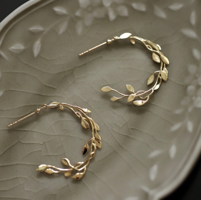 A pair of 925 Sterling Silver Leafy Hoop Earrings with gold plating, displayed on a decorative plate to showcase the intricate leafy branch design and cubic zirconia details.