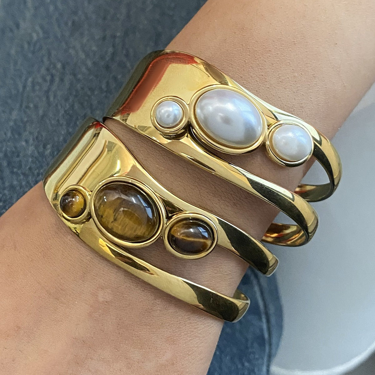 Close-up of a gold-finished stainless steel cuff bracelet set, featuring one with a natural pearl and another with a tiger-eye stone, perfect as a Valentine's gift.